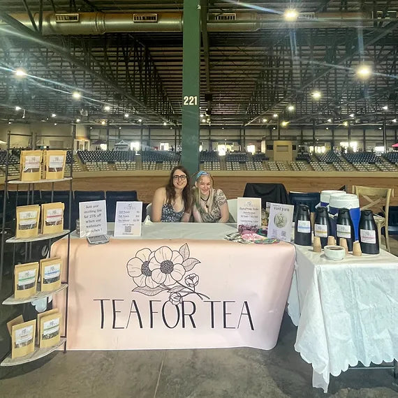 A picture of Ella and Fini, the two Tea For Tea owners, sitting behind a set-up Tea For Tea table at a local fair