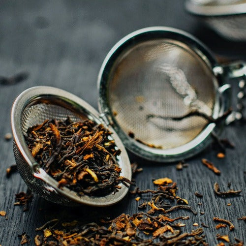 A picture of a metal tea strainer filled with a vibrant black tea with orange leaves in it, sitting on a dark wooden surface